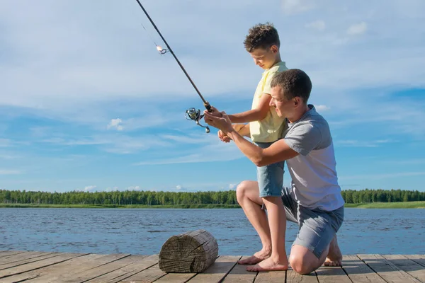 父は美しい風景に対して 魚をキャッチするために釣り竿を保持する方法を桟橋に彼の息子を示しています — ストック写真