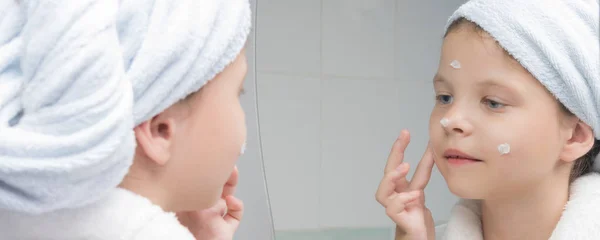 Девушка Белом Халате Полотенцем Голове Увлажняет Лицо Кремом Перед Зеркалом — стоковое фото