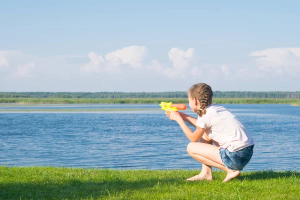 女の子は緑の草の上に座って 水ピストルで遊んで 青い空と湖に対して 碑文のための場所があります — ストック写真