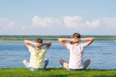 bir erkek ve bir kız yeşil çim enseüzerinde oturuyor ve yoga yapıyor, başlarının arkasında ellerini tutarak, arka görüş, güzel bir manzara fonunda