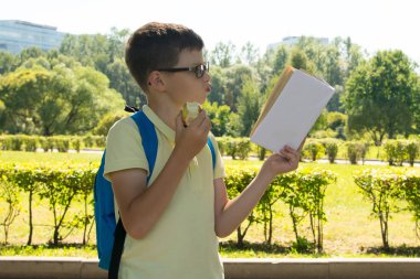 parkta, temiz havada, gözlüklü ve sırt çantası olan çocuk, hevesle bir kitap okuyup elma yiyor.