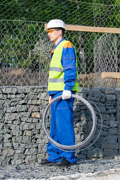 worker in a yellow vest and a white helmet, carries a roll of wire in his hand