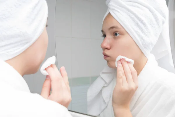 teenager girl wipes her face with acne in the morning, standing in front of a mirror