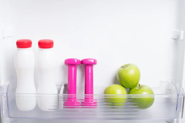 close-up of the shelf in the door of the white refrigerator, green apples, kefir and pink dumbbells for sports