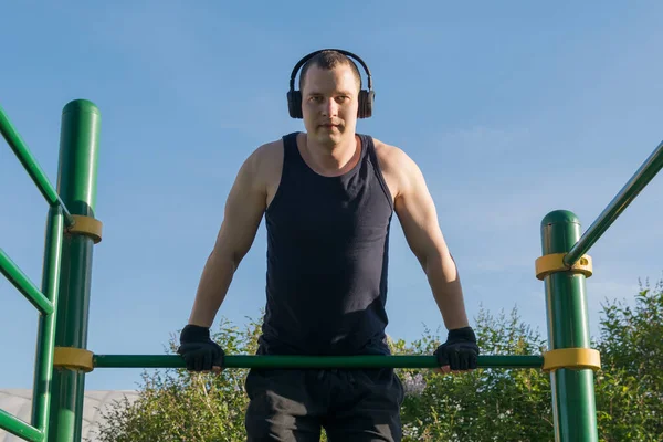 a man goes in for sports on the horizontal bar listening to music on headphones on the street in fine weather