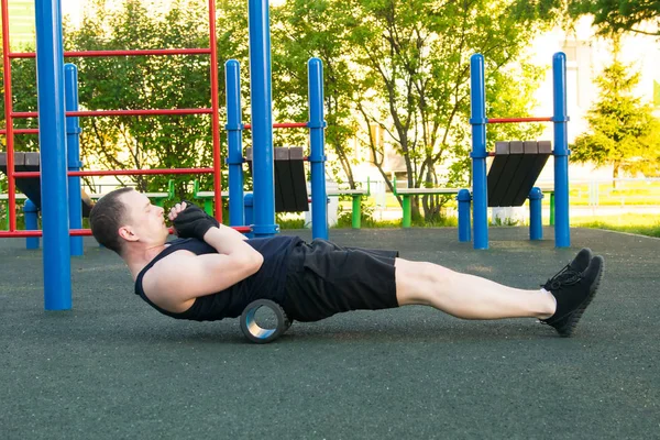 man in black gloves and sneakers, outdoors, doing sports and doing back massage, roller