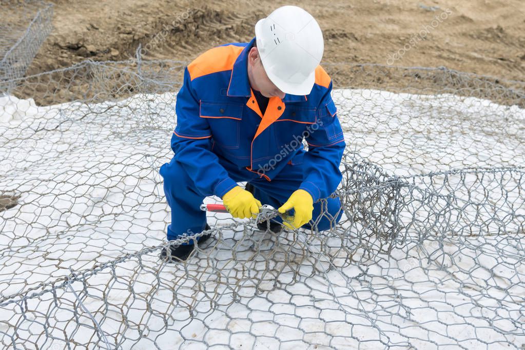 worker in a white helmet and overalls knits a steel wire structure