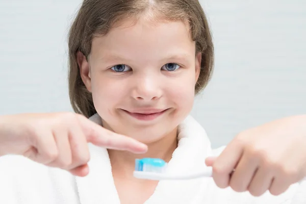 girl in a white coat holds a brush with toothpaste and points a finger at it