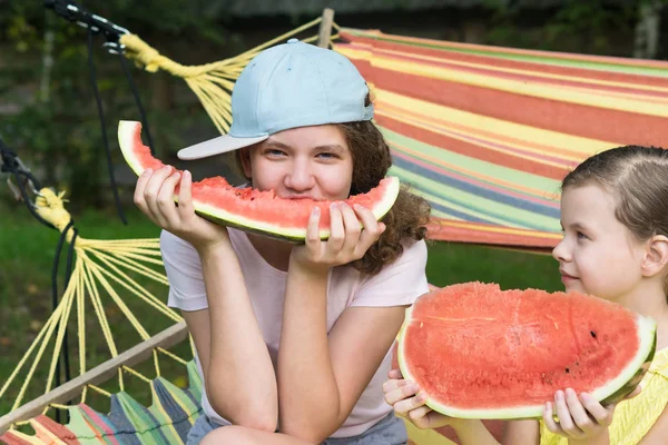 two girls sit in a hammock and eat big red slices of watermelon