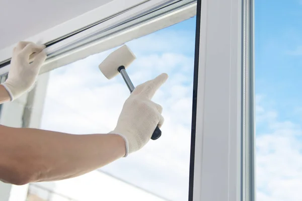 masters hand in protective gloves, fixes a double-glazed window with a plastic baseboard, hammering it with a rubber mallet