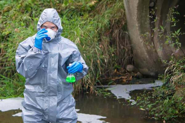 a man in a protective suit, adjusts the mask on the background of drainpipes, looking forward, holding a tube of liquid