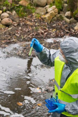 Kazayı tasfiye eden, koruyucu bir giysi ve sarı bir yelek içinde göletin arka planındaki test tüpündeki reaksiyona bakıyor.