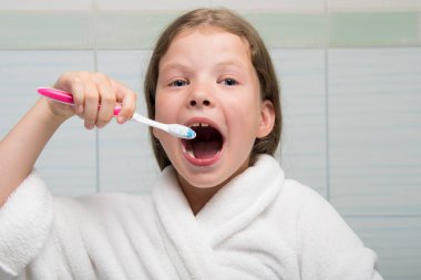 Ağzı açık ve elinde diş fırçası olan bir kız banyoda beyaz bornozla eğleniyor.