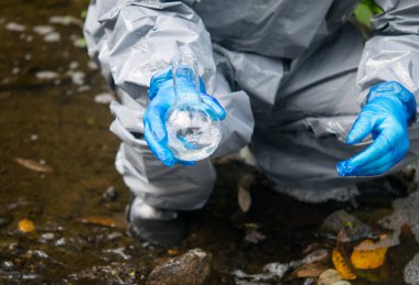 Bilim adamı, koruyucu giysi ve eldiven giymiş, örnek için bir tüp su tutuyor.