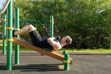 Siyah kolsuz tişörtlü ve şortlu bir adam parkta eğimli bir bankta karın kasları egzersizi yapıyor.