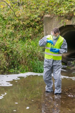 Koruyucu giysili bir adam ve gözlüklü bir solunum cihazı elinde deney tüpleriyle nehrin elverişsiz bölgesinde açık havada test örnekleri tutuyor.
