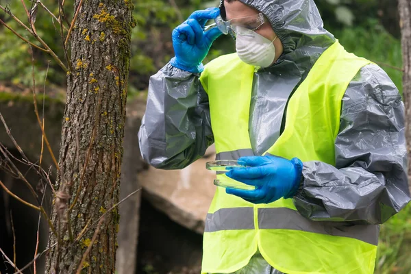 a man in a protective suit and a respirator looks while taking off his glasses on a test tube with a sample of soil taken for research