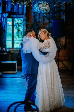 A bride and groom share a romantic moment during their first dance at a warmly lit rustic wedding venue, surrounded by floral dcor and soft blue lighting