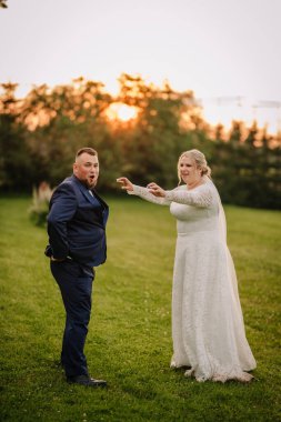 Bride and groom having fun outdoors during sunset. The groom poses playfully while the bride reaches toward him, creating a candid and joyful wedding moment.