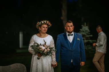 Cesis, Latvia - July 17, 2025 - A bride and groom stand side by side at night, holding hands. The bride wears a lace gown with a floral crown and bouquet, while the groom is in a blue suit and bow tie.