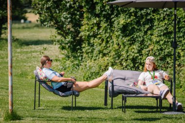 Sigulda, Letonya - 18 Temmuz 2025 - Çimenli bir açık hava hippi temalı etkinliğinde iki kadın, huzurlu bir parkta büyük bir şemsiye altında içki ve güneşin tadını çıkarıyor..