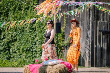 Sigulda, Letonya - 18 Temmuz 2025 - Renkli, hippi esinlenilmiş iki kadın açık hava festivalinde saman balyasının önünden geçtiler. Tanımlanabilir kişilere bağlı editoryal kullanım.