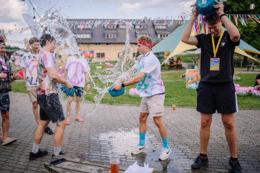 Sigulda, Letonya - 18 Temmuz 2025 - Bir hippi festivalinde bir grup insan kovaları kullanarak birbirlerine su sıçratıyorlar. Hareket enerji, kahkaha ve yaz atmosferiyle dolu..