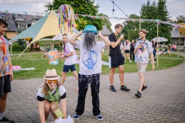 Sigulda, Letonya - 18 Temmuz 2025 - Bir hippi festivalinde bir adam eğlence olsun diye kafasına kova döker, diğerleri de seyreder ve güler. Sahne renkli, neşeli ve yaz enerjisiyle dolu..