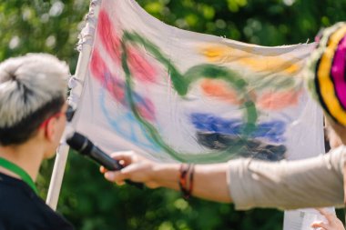 Sigulda, Letonya - 18 Temmuz 2025 - Bir hippi festivalinde bir insan soyut şekilli renkli bir bayrak tutarken, diğeri mikrofona konuşuyor. Canlı bir ortamda yaratıcı ifade.