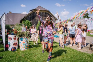 Sigulda, Letonya - 18 Temmuz 2025 - Canlı hippi kıyafetleri giymiş bir adam güneşli bir gökyüzünün altında insanlarla çevrili canlı bir açık hava festivalinde barış işareti gösteriyor..