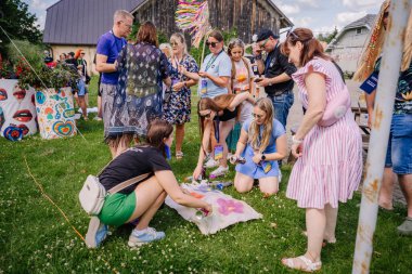 Sigulda, Letonya - 18 Temmuz 2025 - Bir hippi festivalinde insanlar renkli sprey boyalarla kumaş süslemek için çimlerin üzerinde toplanıyor, canlı psikedelik sanatın yanında yaratıcı bir grup aktivitesinin keyfini çıkarıyorlar..