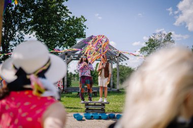 Sigulda, Letonya - 18 Temmuz 2025 - Açık hava festivalinde renkli kurdeleler, yaz dekorları ve güneşli günün tadını çıkaran bir seyirci ile konuşan iki hippi kıyafeti giymiş iki kişi.
