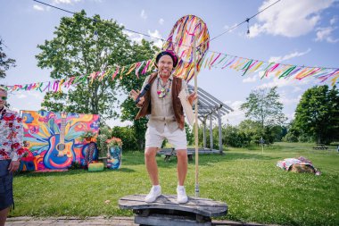 Sigulda, Letonya - 18 Temmuz 2025 - Hippi kıyafeti giymiş bir adam mikrofonlu bir bankta duruyor, renkli bir açık hava festivalinde gülüyor ve kurdele ve sanatla süslenmiş poz veriyor. Hippi..