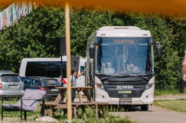 Sigulda, Letonya - 18 Temmuz 2025 - Beyaz bir İskandinav otobüsü, park halindeki arabalar ve piknik mobilyalarıyla çevrili, açık hava hippi tarzı bir festivalin önüne park edilmişti. Sadece editör kullanımı.