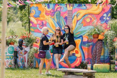 Sigulda, Letonya - 18 Temmuz 2025 - Üç kadın açık hava festivalinde psikedelik sanat ve mankenlerle canlı, hippi tarzı bir arkaplan önünde mutlu poz veriyor. Tanımlanabilir kişilere bağlı editoryal kullanım.