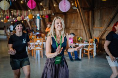 Sigulda, Letonya - 18 Temmuz 2025 Hippi temalı bir festivalde gülümseyen kadınlar, kırsal bir ahşap mekanda renkli asılı dekorasyonlarla çevrili, müzikten ve iyi titreşimlerden keyif alan, iç mekanlarda dans ediyorlar..