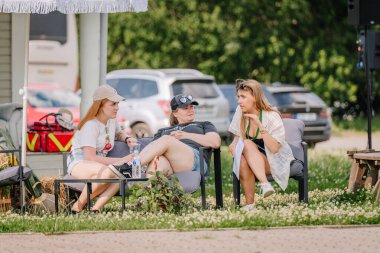 Sigulda, Letonya - 18 Temmuz 2025 - Üç kadın rahat bir yaz atmosferinde çimenler, bitkiler ve park halindeki arabalarla çevrili hippi temalı bir açık hava etkinliği sırasında şemsiye altında oturup konuşur..