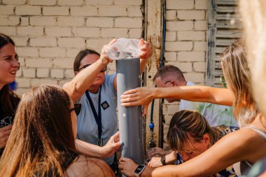 Sigulda, Letonya - 18 Temmuz 2025 - Bir grup insan, çoğunlukla kadınlar, bir boruya su dökülmesini içeren açık hava hippi tarzı bir yarışmada işbirliği yapıyorlar. Takım ruhu ve odaklanması görünür..