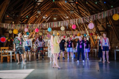 Sigulda, Letonya - 18 Temmuz 2025 - Bir grup insan, renkli fenerler ve kurdelelerle süslenmiş ve çıplak ayaklı bir hoparlörü dikkatle dinleyen kapalı mekan temalı bir atölyede veya toplantıda.