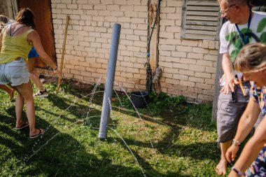 Sigulda, Letonya - 18 Temmuz 2025 - Hippi temalı açık hava grubu aktivitesi katılımcılar eğlenceli bir su borusu yarışması yapıyor, güneşli bir günde gülüyor ve ıslanıyorlar. Saf yaz neşesi ve takım çalışması..