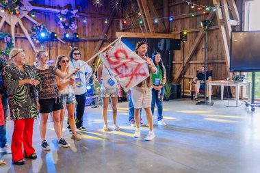 Sigulda, Letonya - 18 Temmuz 2025 - Hippi temalı kapalı mekan festivalinde renkli bir barış bayrağı taşıyan bir grup insan, dekoratif ışıklar ve ahşap kirişler altında eğleniyor ve gülümsüyor..