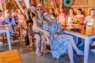 Sigulda, Letonya - 18 Temmuz 2025 - Bir grup gülen insan, kapalı mekan temalı bir etkinlikte renkli kıyafetler ve çiçek desenleri giyerek atmosferin tadını çıkarıyor ve dekore edilmiş masalarda oturuyorlar. Hippi..