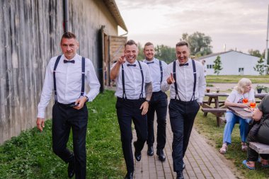 Sigulda, Letonya - 18 Temmuz 2025 - Pantolon askısı ve papyonlu dört adam güvenli bir şekilde dışarıda yürüyor, gülümsüyor ve neşeyle işaret ediyor. Hippi temalı değil.