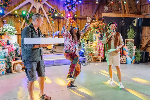 Sigulda, Letonya - 18 Temmuz 2025 - Yama pantolonlu bir adamla sahnede neşeyle dans eden, içki içen diğerleri renkli ışıklar altında hediyelerle neşelenirken hippi temalı festival kutlaması.