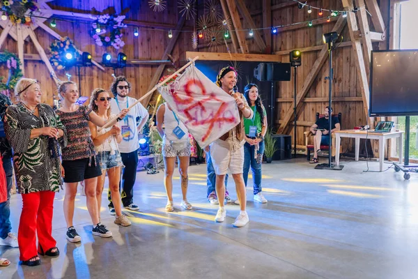 Sigulda, Letonya - 18 Temmuz 2025 - Hippi temalı kapalı mekan festivalinde renkli bir barış bayrağı taşıyan bir grup insan, dekoratif ışıklar ve ahşap kirişler altında eğleniyor ve gülümsüyor..