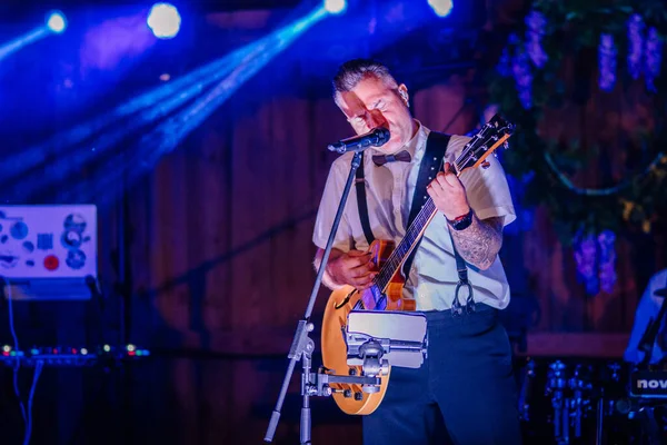 Sigulda, Letonya - 18 Temmuz 2025 - Gitarist hippi festivali sırasında kırsal alanda dramatik bir mavi ışıklandırma altında papyon ve pantolon askısı takarak sahnede tutkuyla şarkı söylüyor..