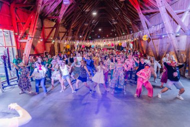 Sigulda, Letonya - 18 Temmuz 2025 - Hippi kostümlü büyük bir grup, renkli ışıklar ve fenerlerle dekore edilmiş bir ahırda retro temalı bir etkinlik sırasında enerjik bir şekilde dans ediyorlar..