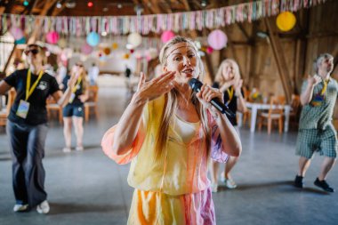 Sigulda, Letonya - 18 Temmuz 2025 - Renkli hippi kıyafetli gülümseyen kadın arka planda insanlar, şenlikli süslemeler ve asılı fenerler altında eğlenceli bir dans etkinliği düzenliyor..