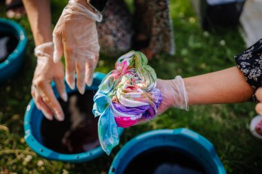 Güneşli bir günde açık havada bir hippi kravat boyama atölyesi sırasında renkli pigmentlere boyanmış spiral bir tişört tutan eldiven giymiş ellerin yakın çekimi..