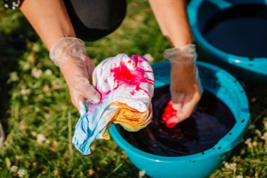 Eldivenli eller, açık hava hippi temalı kravat boyama atölyesi sırasında beyaz gömleğe kırmızı boya sürerler. Renkli kovalar ve parlak renk lekeleri vardır..